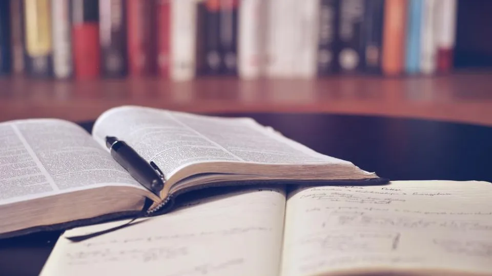 Open books and a pen on a table in front of a bookshelf