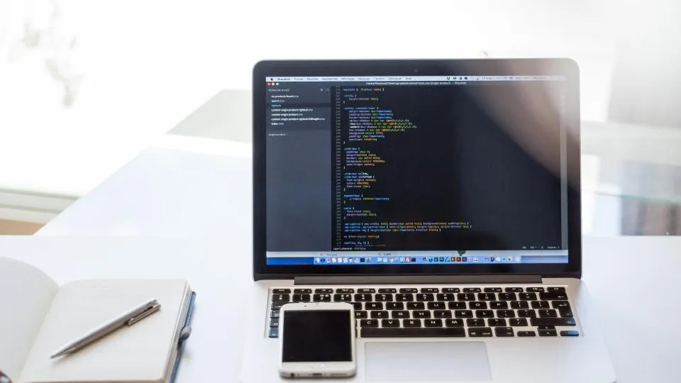 A laptop showing code on a bright desk, with a notebook and a phone