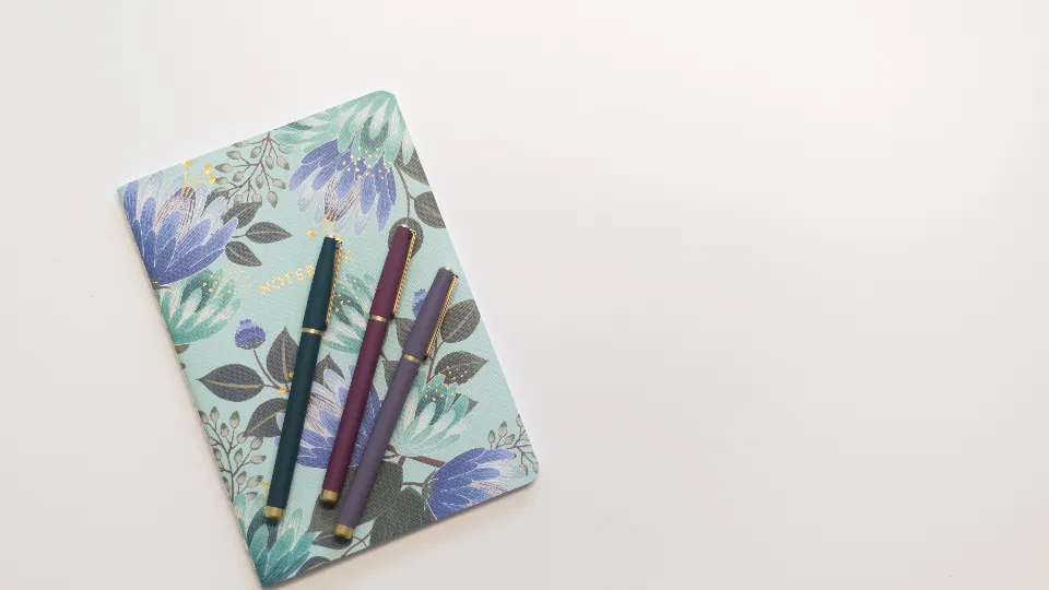 A floral notebook with two pens on a pale desk